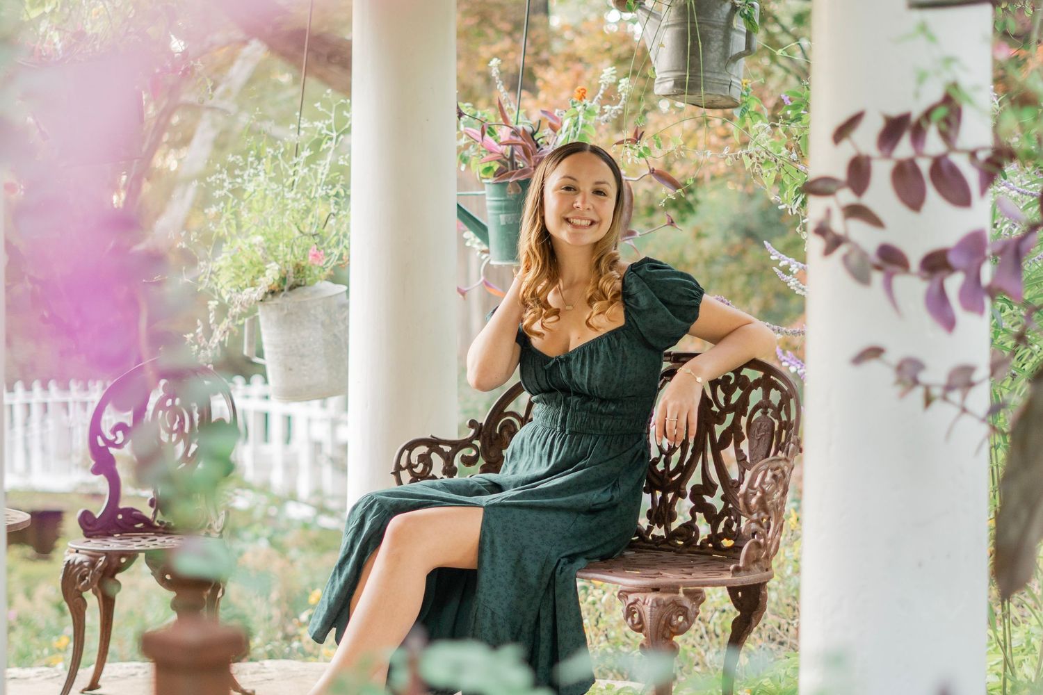 A smiling woman in a dark green dress sits on an ornate bench on a flower-filled garden porch.