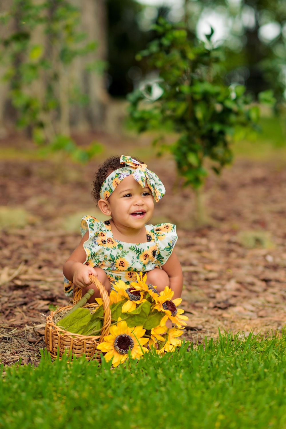 A smiling baby in a floral outfit sits with sunflowers in a basket on grass.