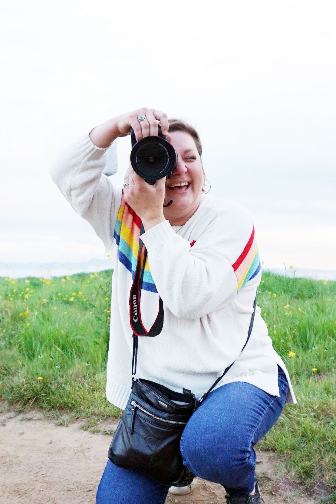 A person in a white sweater with rainbow stripes takes photos in a grassy field on a sunny day.