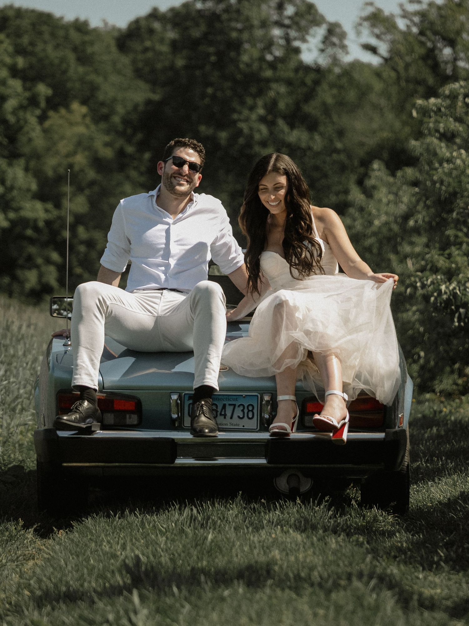 A sunny outdoor portrait of a couple sitting on the back of their vintage blue convertible.