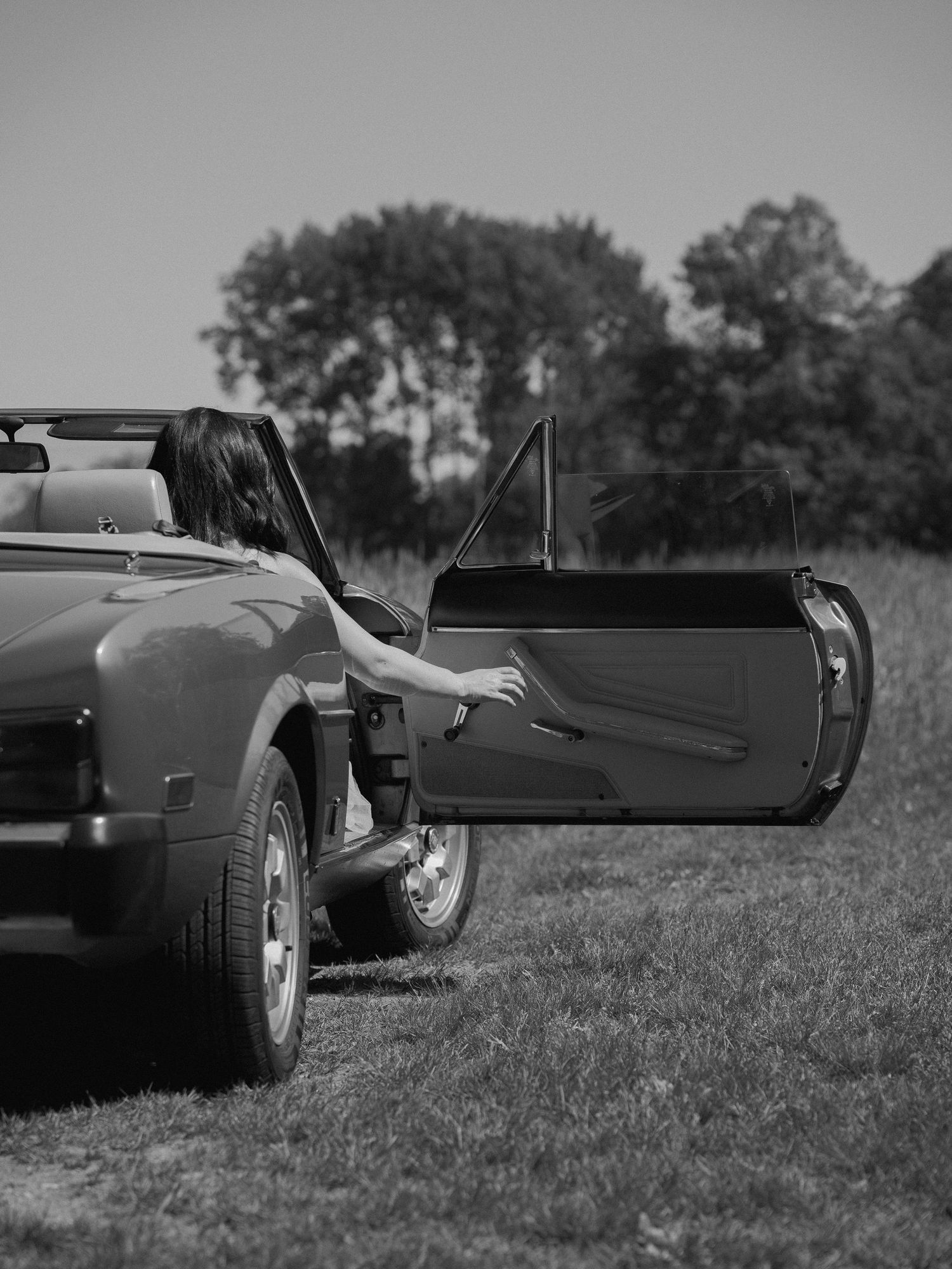 A black and white photo showing the door of a classic convertible car opened in a field.