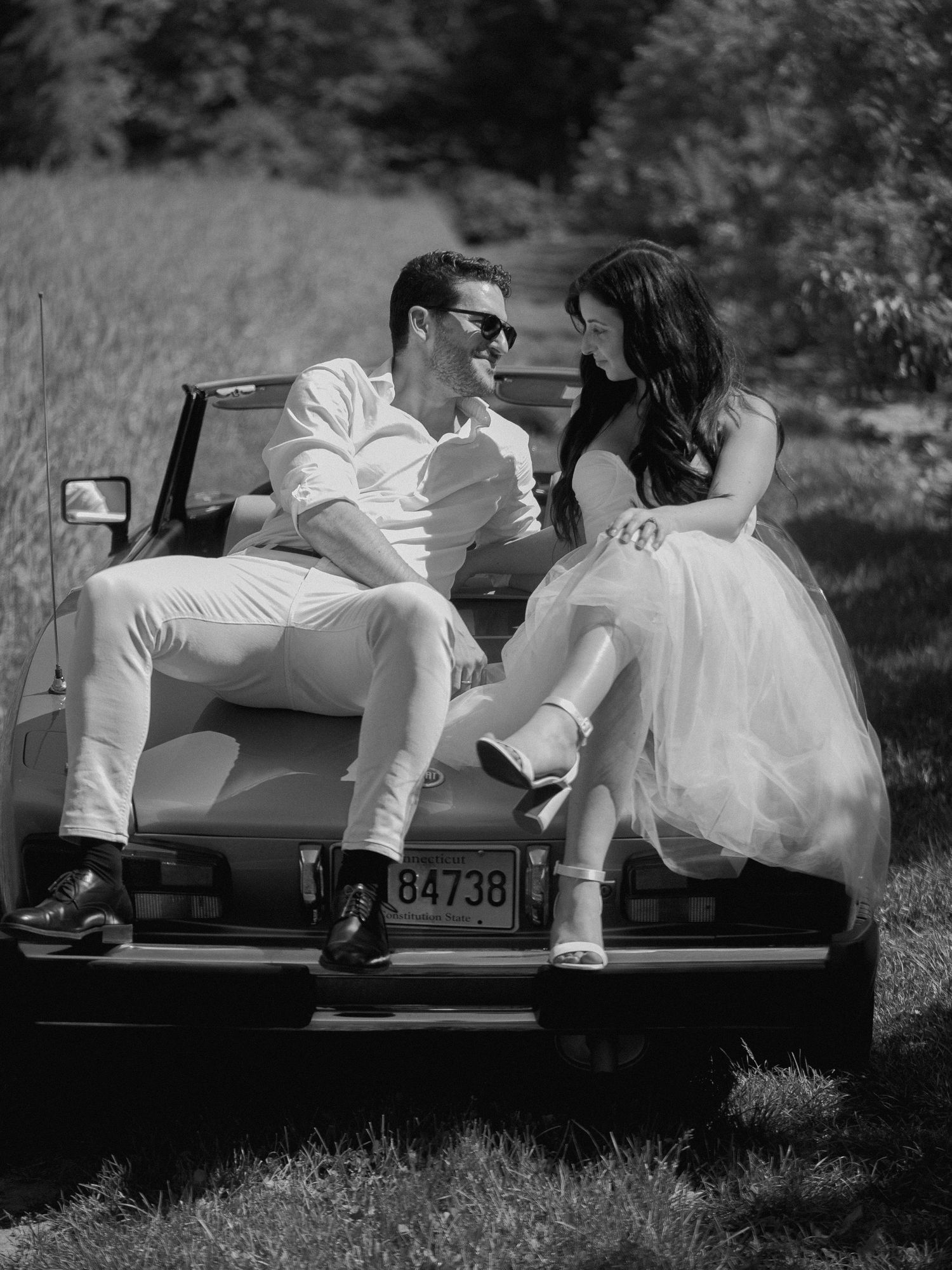 A couple in white outfits sits on the trunk of their vintage car during a countryside photoshoot.