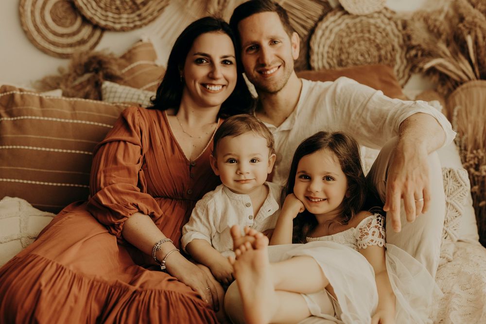 Smiling family of four with two young children cuddle together on a cozy boho couch with woven wall decor.