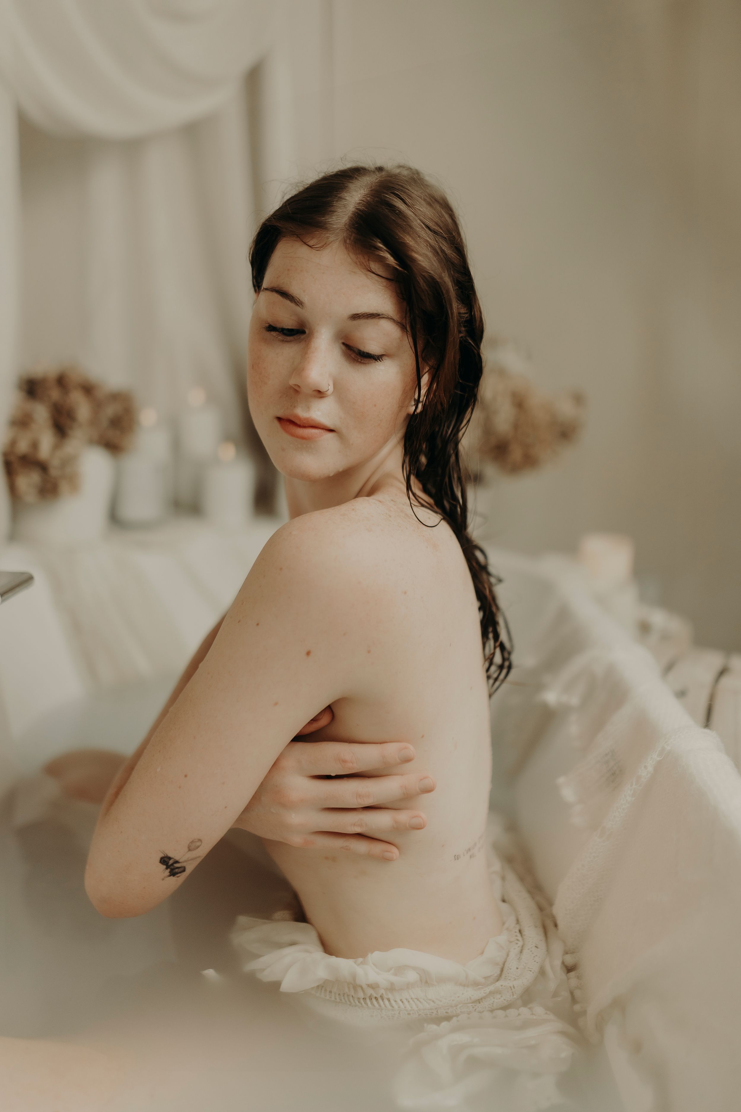 Woman with wet hair and closed eyes sits in a bath with her arm across her chest, surrounded by candles and dried flowers.