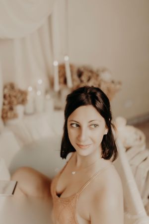 Dark-haired woman in a peach lace camisole looks aside with a gentle smile beside a candlelit bathtub and dried flowers.