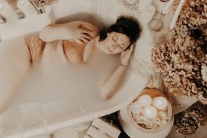 Woman relaxes in a milk bath with her eyes closed, surrounded by candles, dried flowers and a bowl of bath salts.