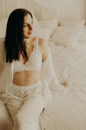 Dark-haired woman in a white lace bralette, loose pants and open shirt sits on a bed and looks to the side in soft light.