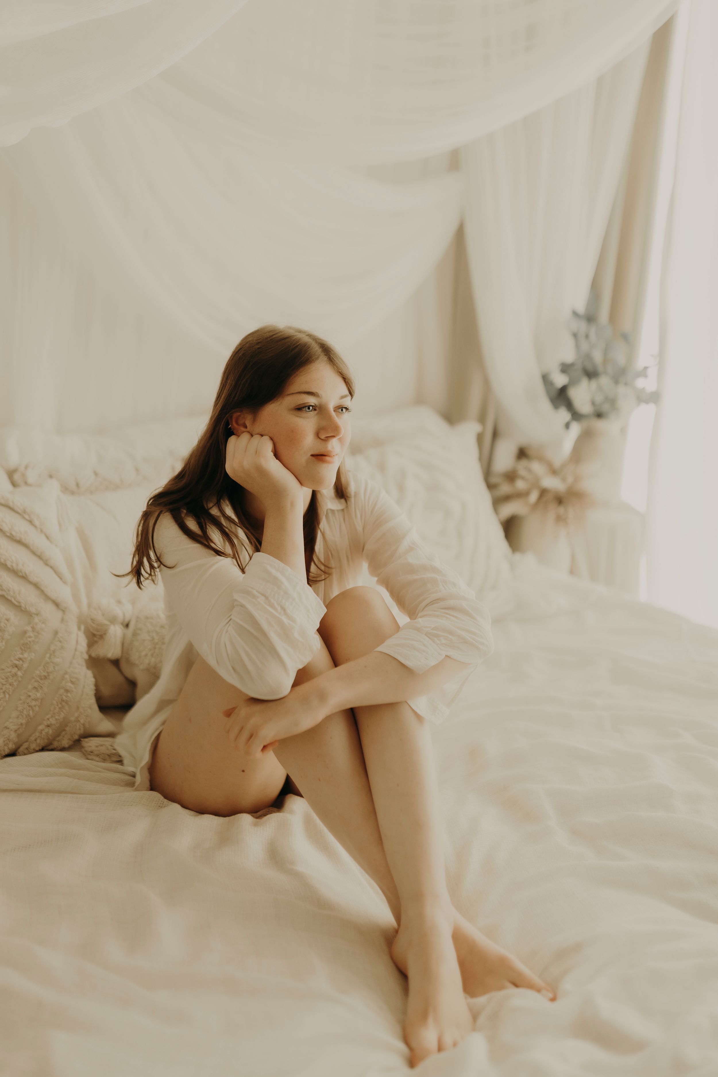 Young woman in a white shirt sits on a bed with sheer canopy curtains, resting her chin on her hand and gazing away.