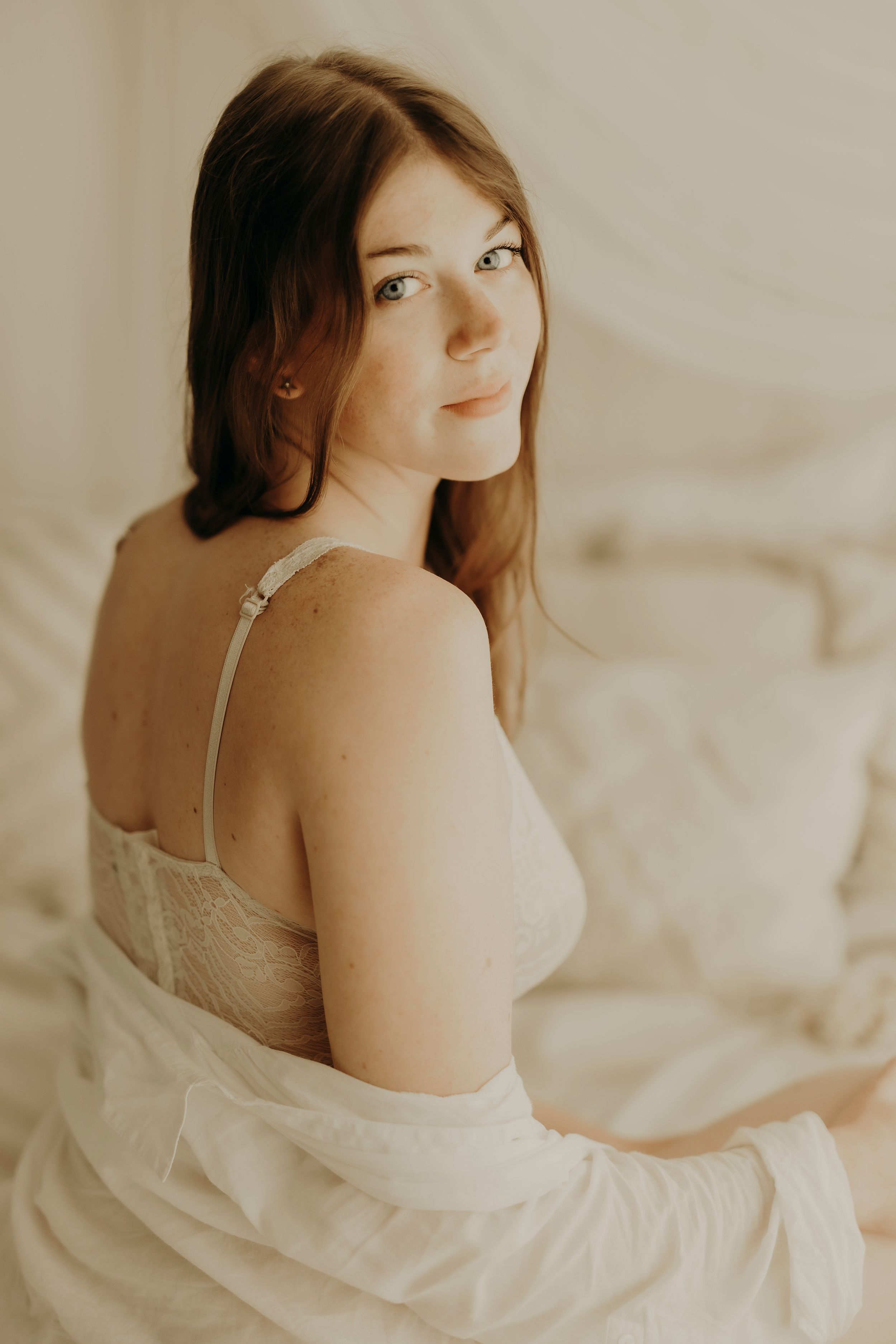 Portrait of a young woman with freckles and long auburn hair glancing over her shoulder in soft light on a white bed.