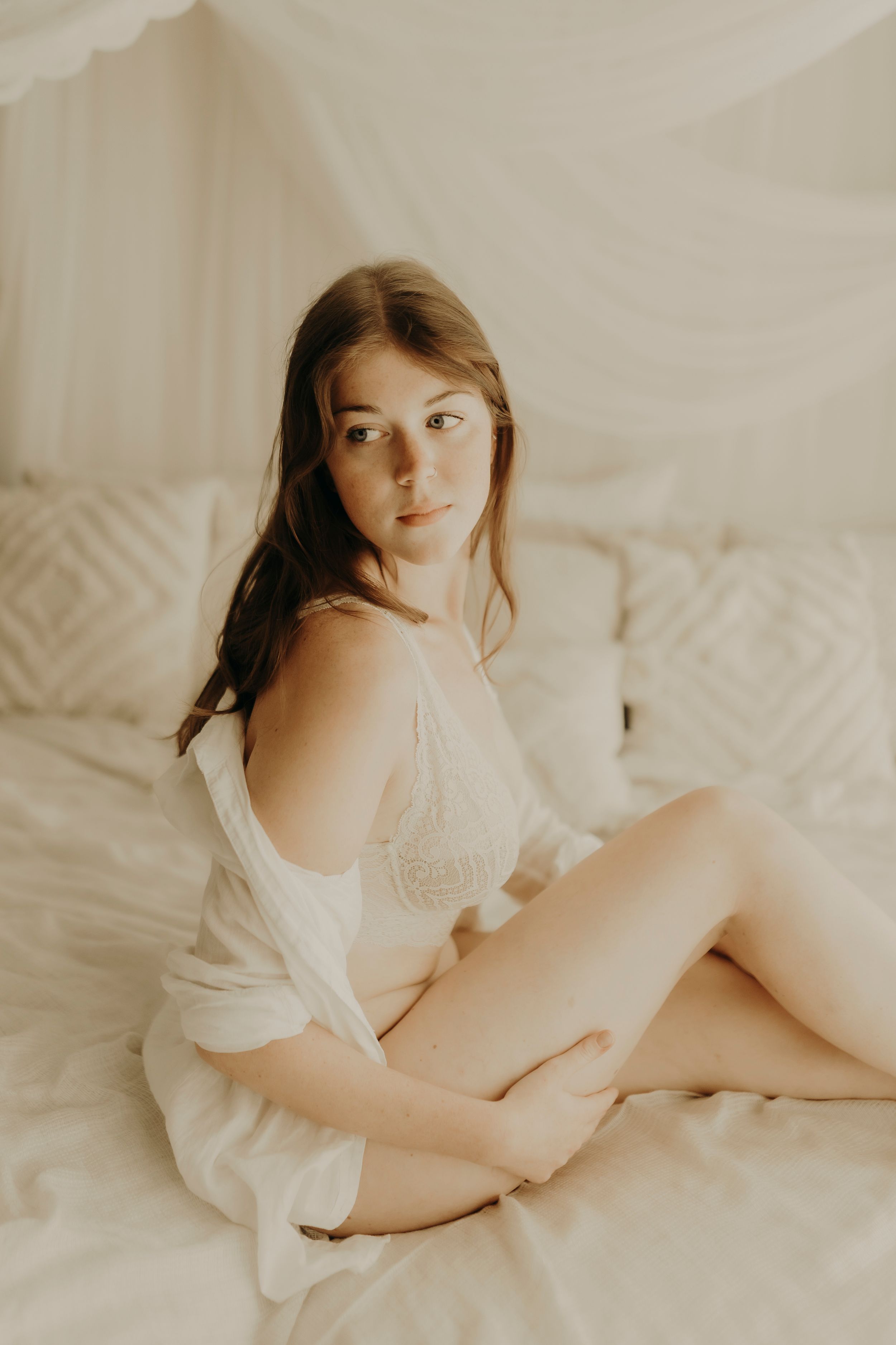 Woman in white lace lingerie and an open shirt sits on a bed with sheer drapes, looking aside in soft natural light.