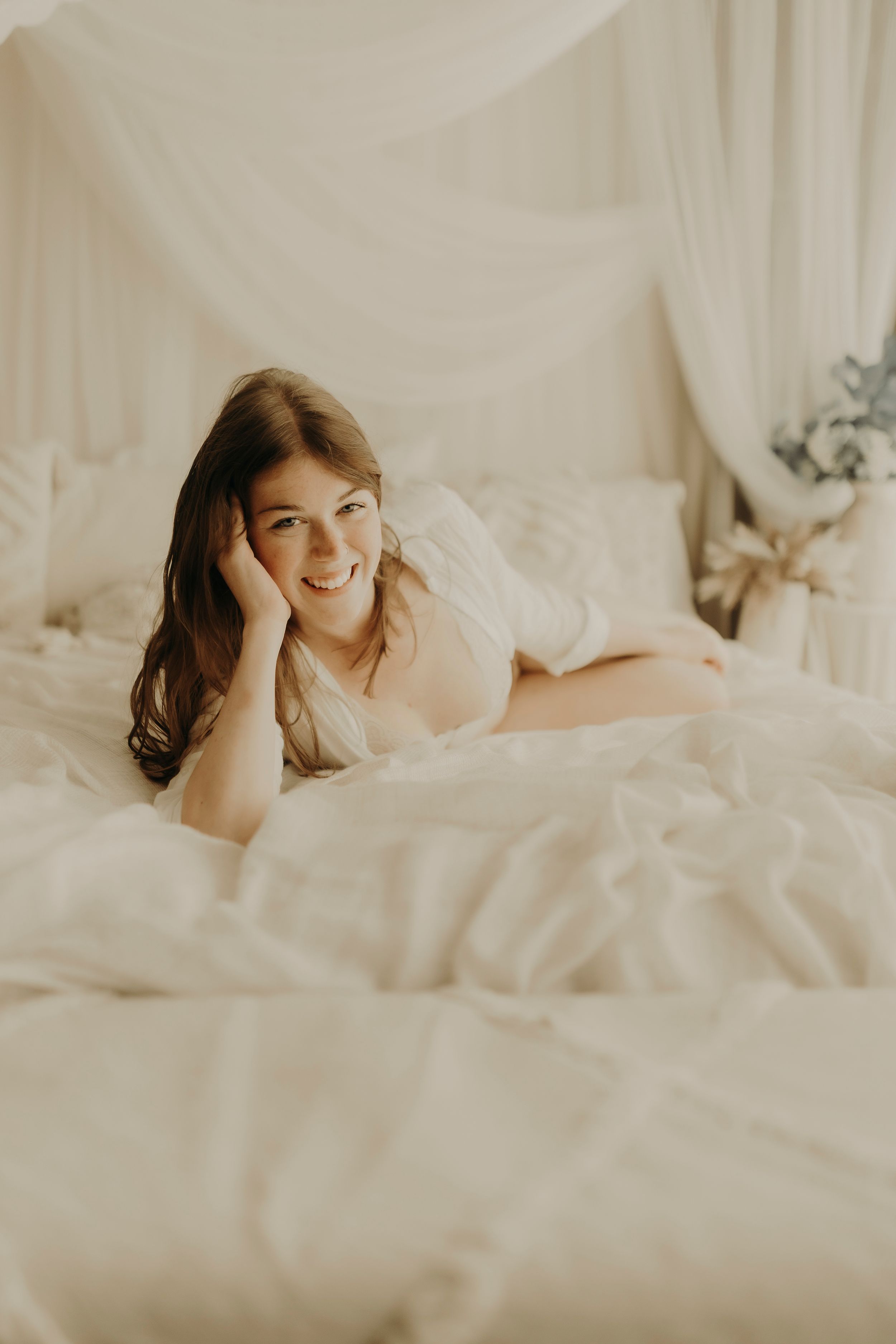 Smiling woman with long wavy hair lies on a white bed propping her head on her hand, with sheer curtains behind her.