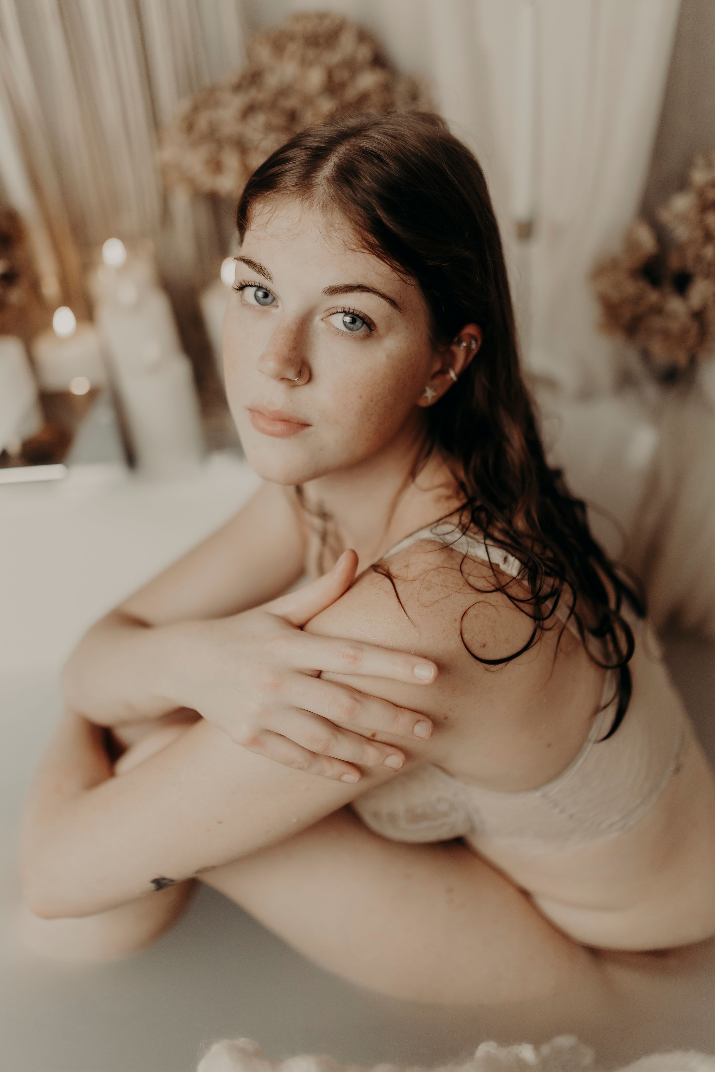 Freckled woman with wet hair sits in a milk bath with dried flowers and candles, hugging her knees and looking at the camera.