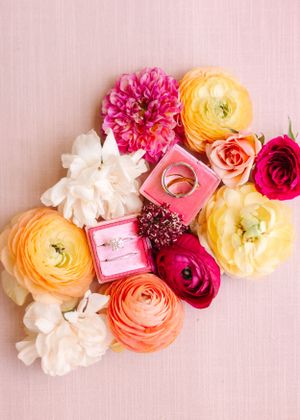 Pink and yellow ranunculus and roses arranged with wedding rings in a pink box on light pink background.