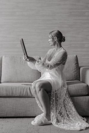 A lace-robed figure sitting on grey couch looking at handheld mirror in black and white photography.