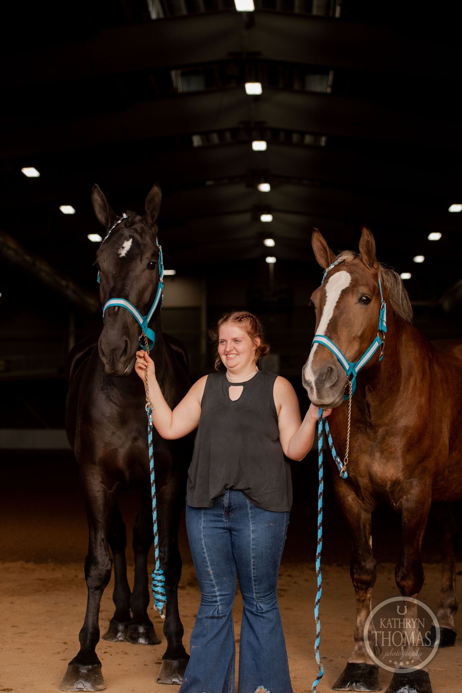 Two horses wearing blue halters stand side by side in a dark indoor arena.