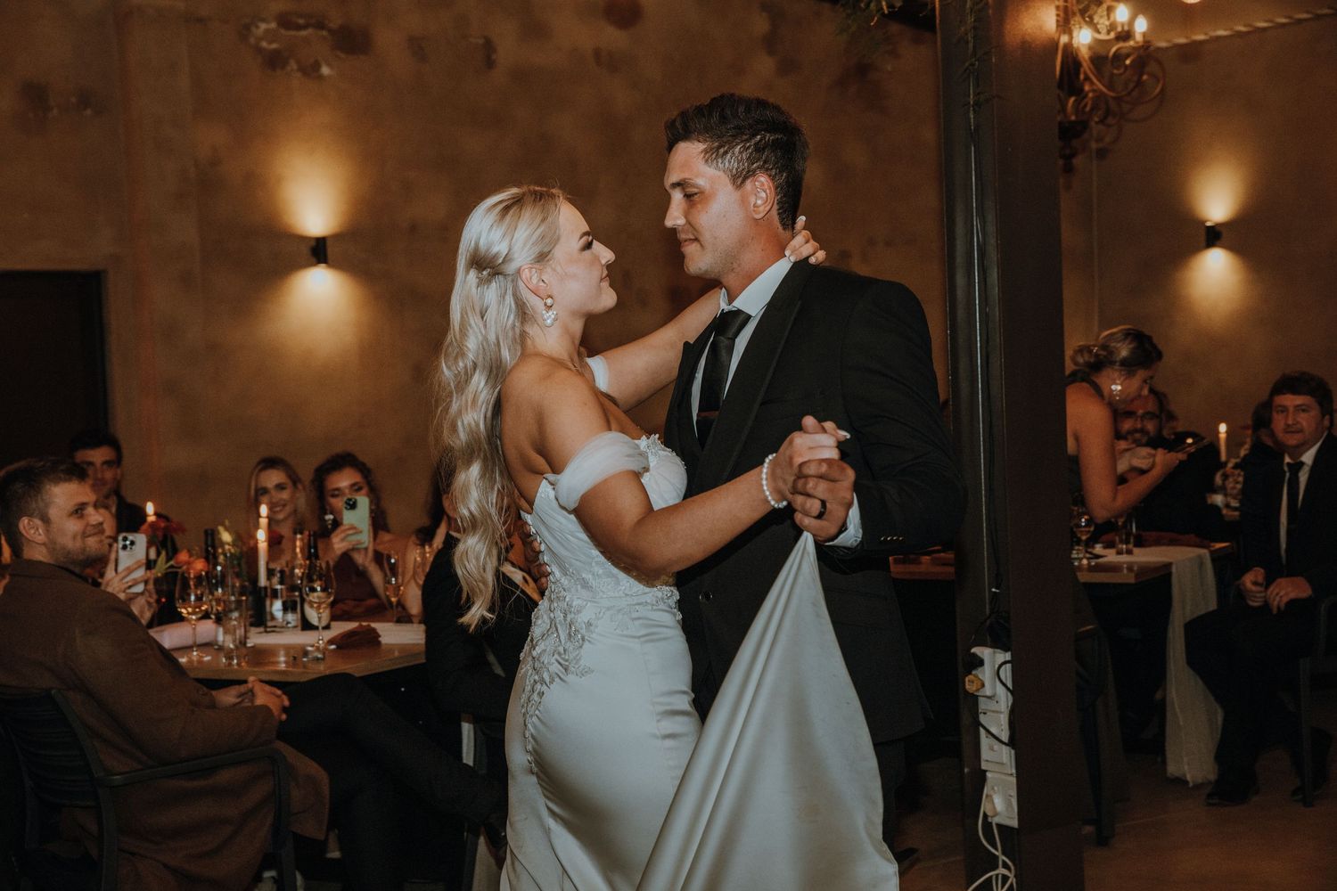 A romantic wedding couple shares their first dance in a warmly lit reception hall with guests watching.