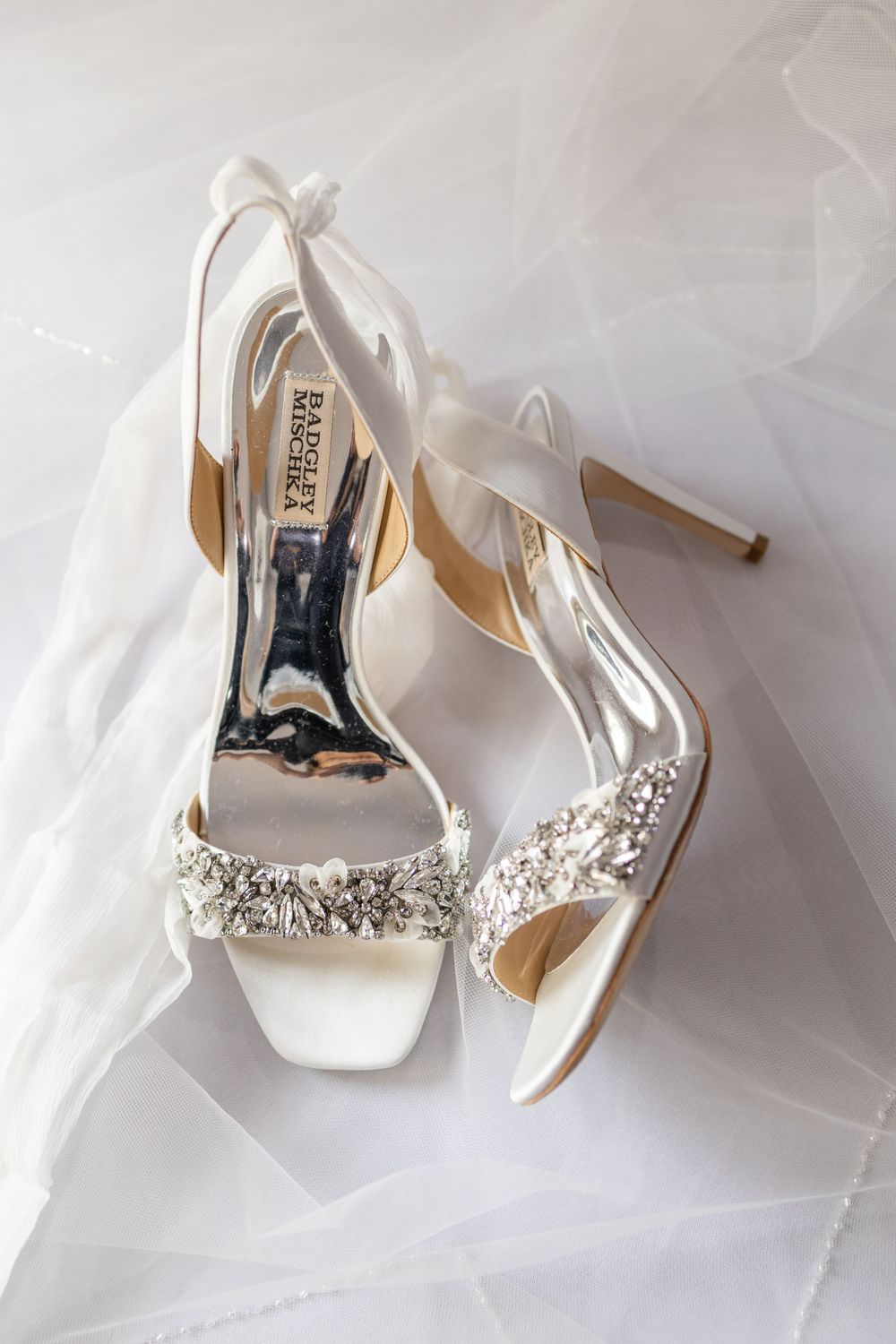 White satin bridal heels with crystal embellishments on tulle backdrop.