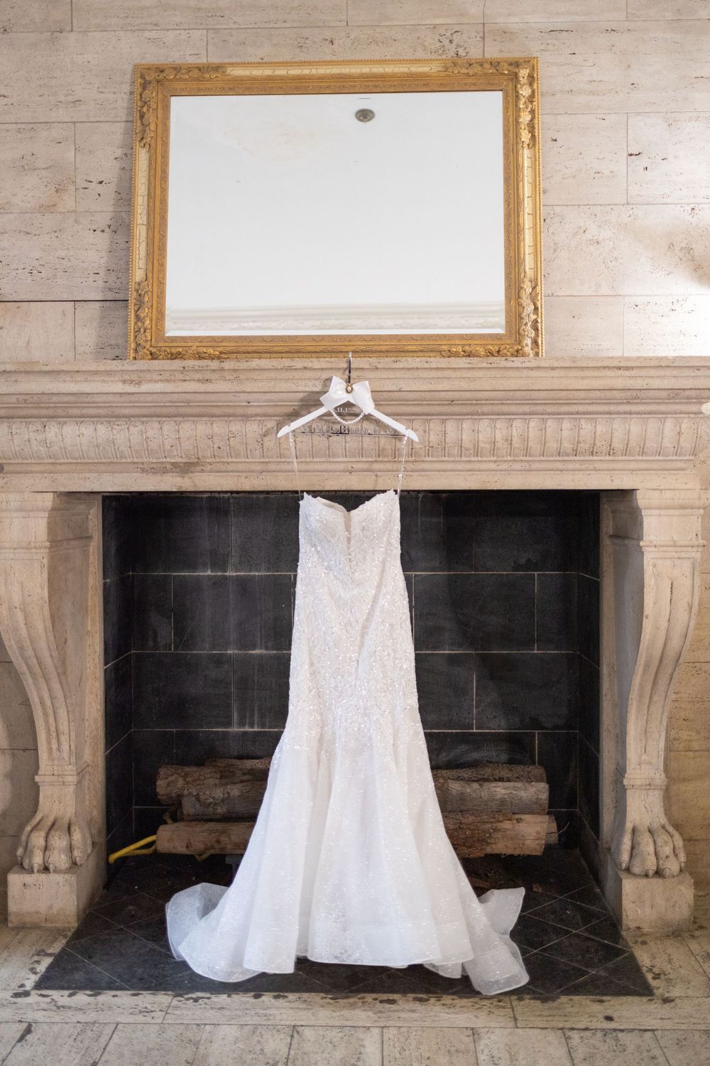 An elegant white wedding dress hanging on display in front of a rustic stone fireplace.