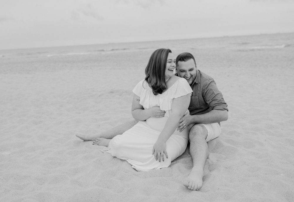 Black and white romantic portrait of couple sitting close together on sandy beach.
