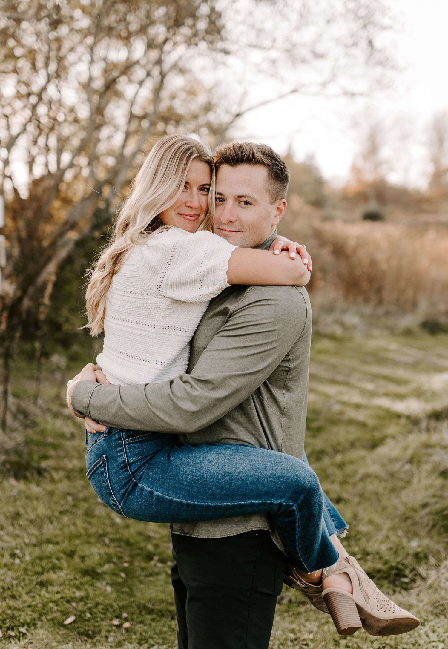 A couple in casual attire shares affectionate embraces in a sunlit outdoor setting with autumn colors.
