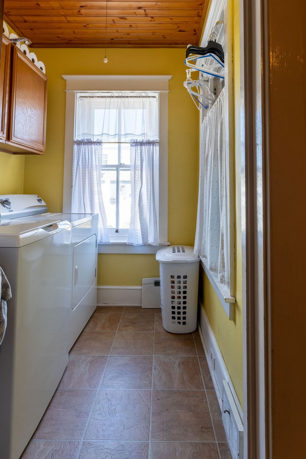 Narrow laundry room with yellow walls, white appliances, and a window with sheer curtains.