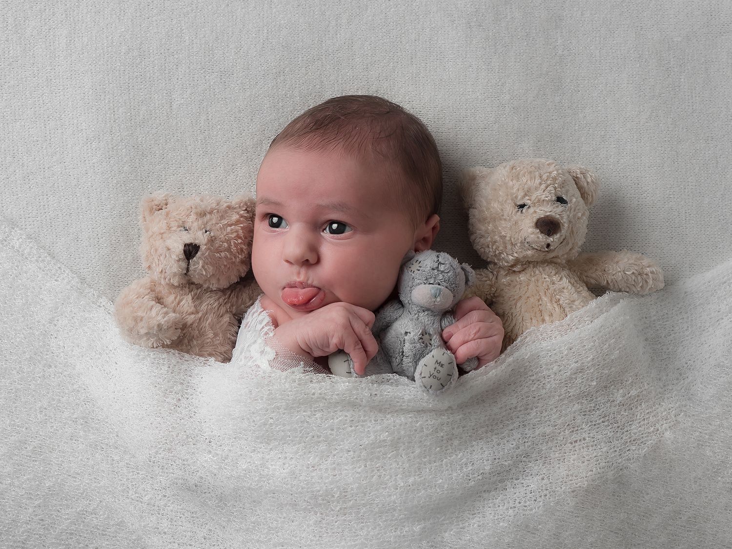 baby sticking out her tongue, tucked in bed with lots of teddies