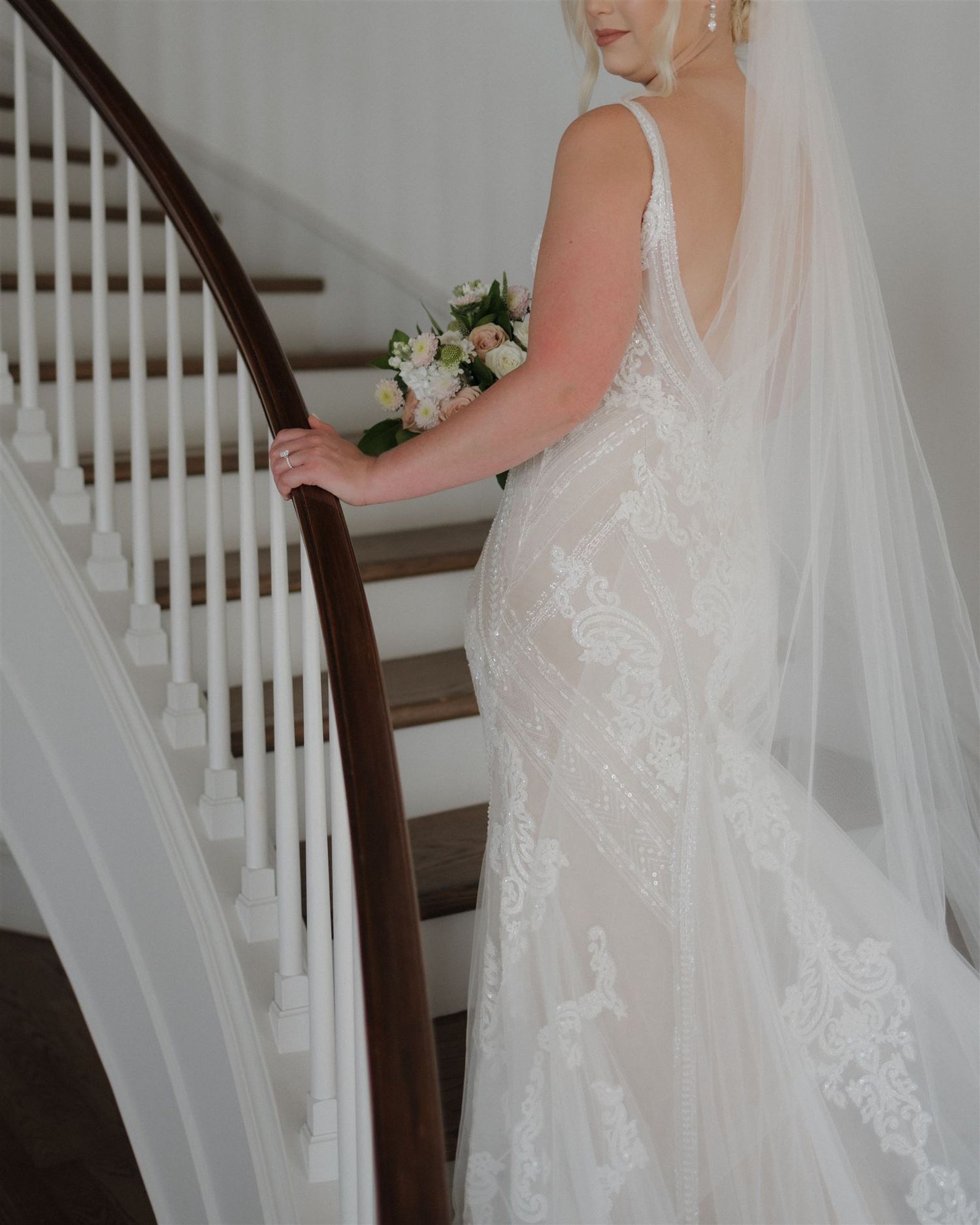 Side view of bride in open-back wedding gown holding bouquet on wooden staircase.