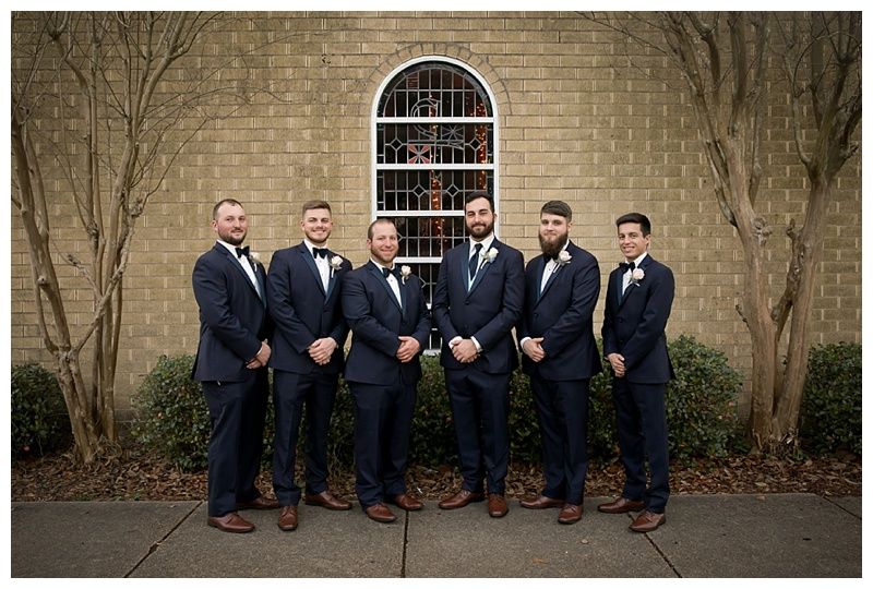 Wedding party posed against brick wall outdoors in formal attire.