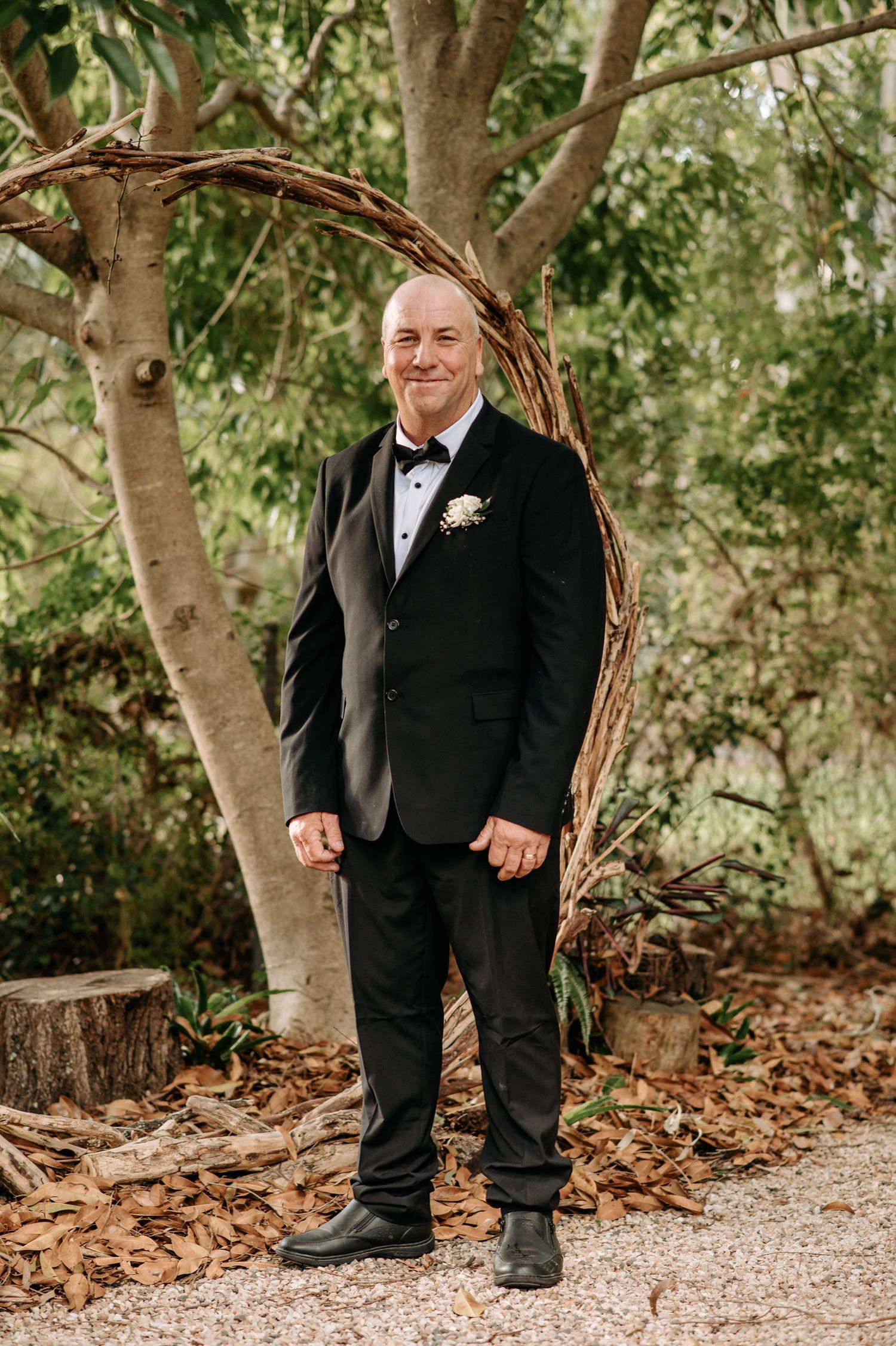 Groom in classic black tuxedo poses against garden backdrop during wedding ceremony.