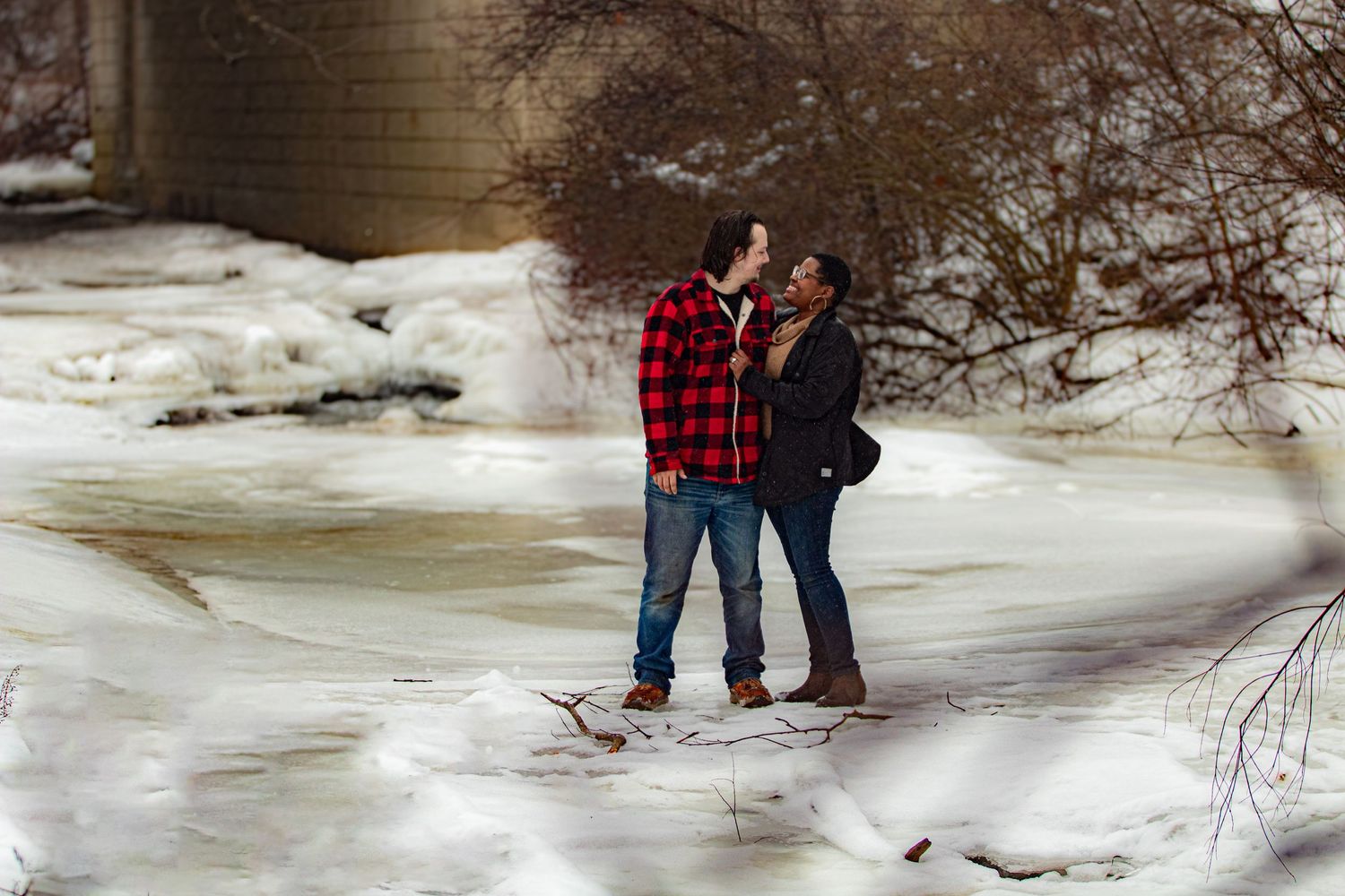An engaged couple laughing to themselves on a frozen riverbed with a small frozen waterfall