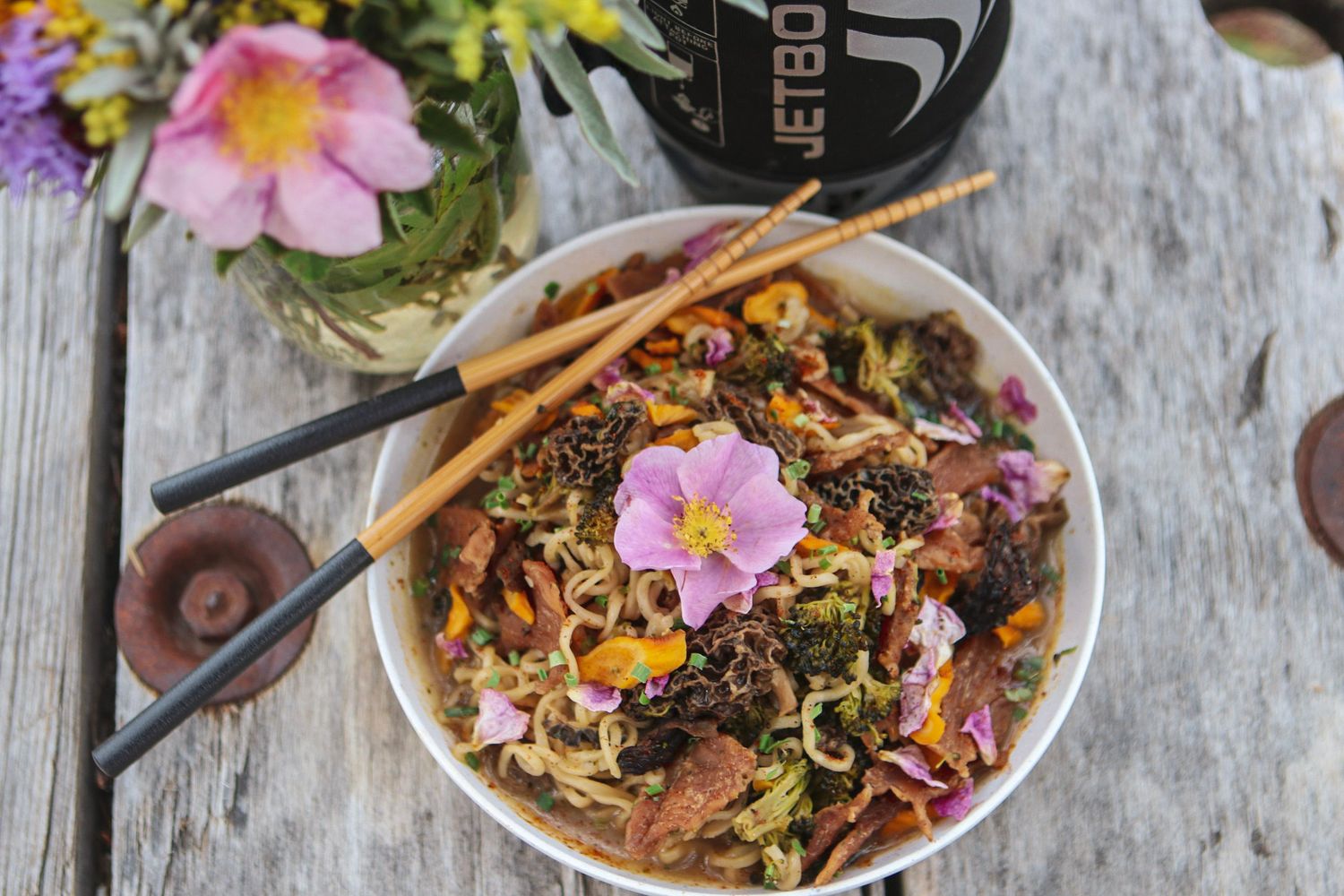 Asian noodle bowl with pink flower garnish and chopsticks on weathered wood table.