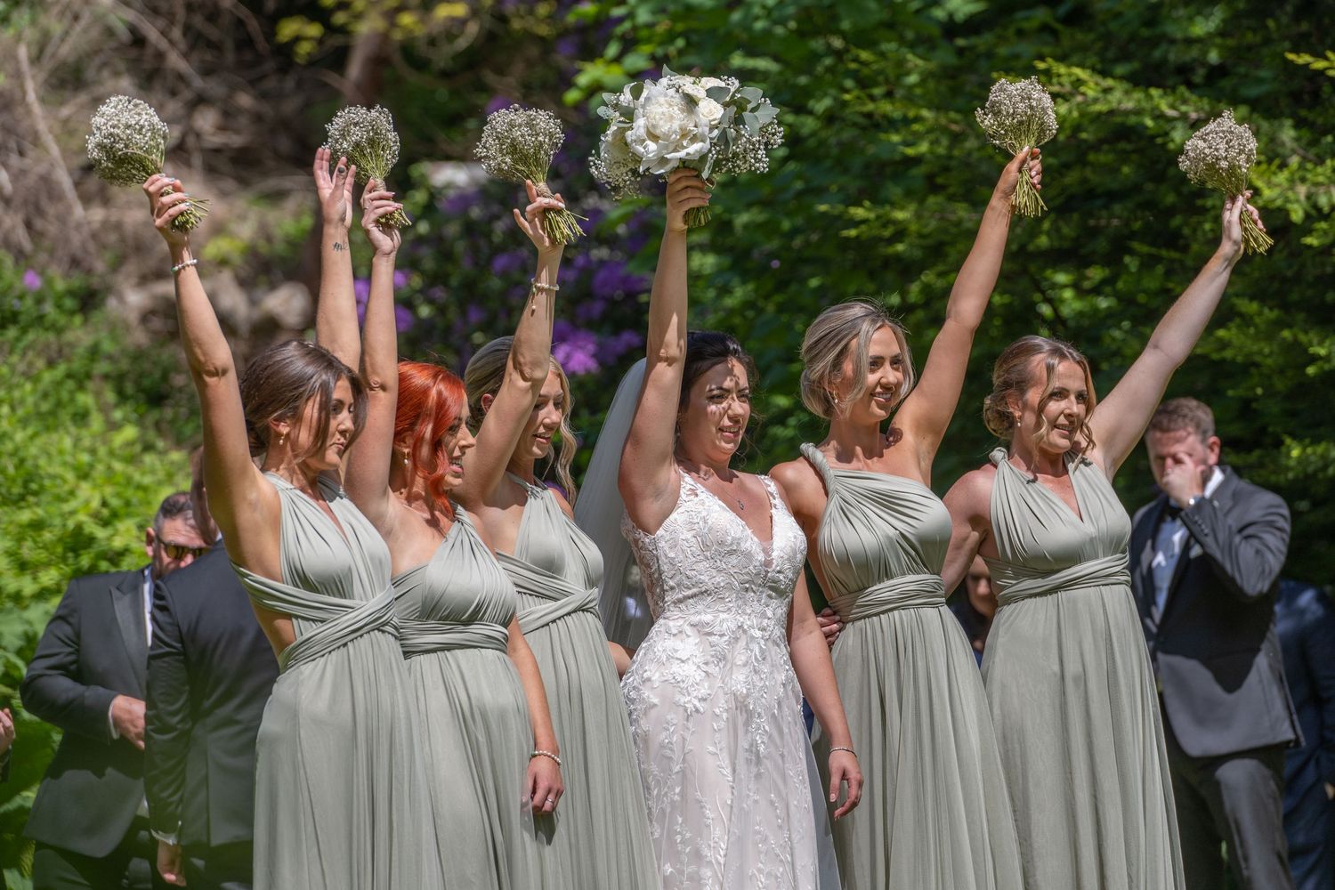 A celebratory group wearing sage green dresses and white holding bouquets up in the air outdoors.