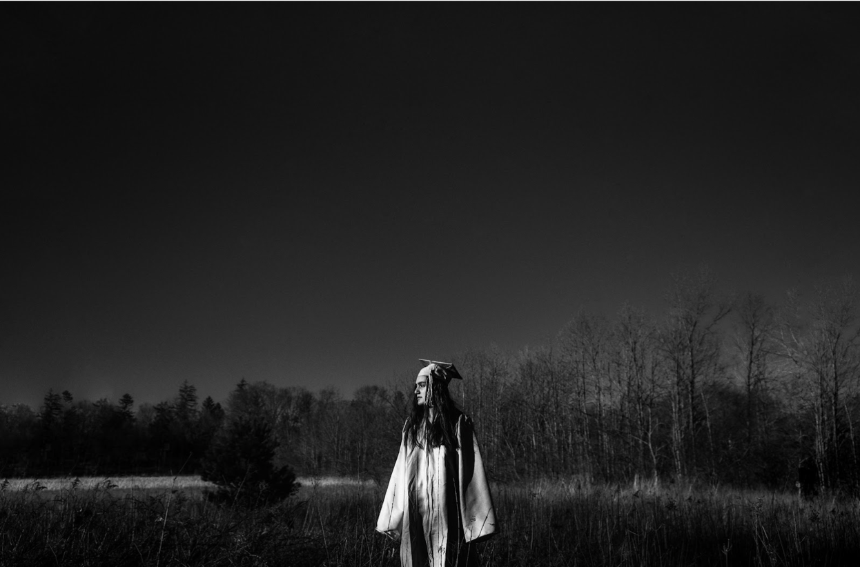 A figure in white stands alone in a dramatic black and white field landscape at dusk.
