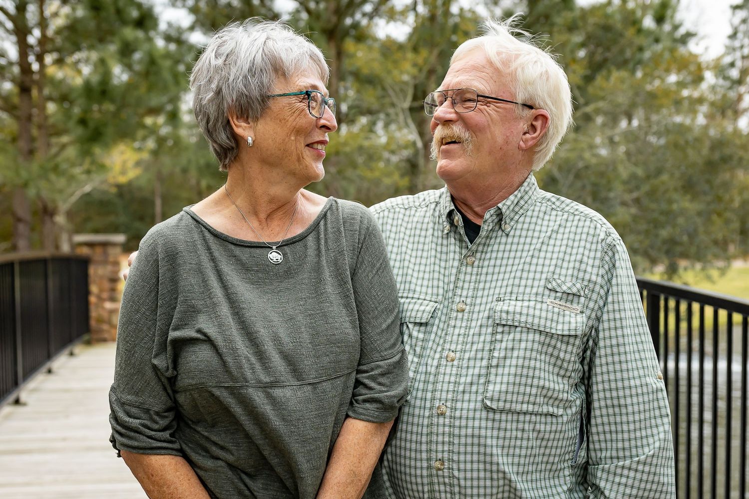 Elderly couple laughing together on wooden deck surrounded by trees.