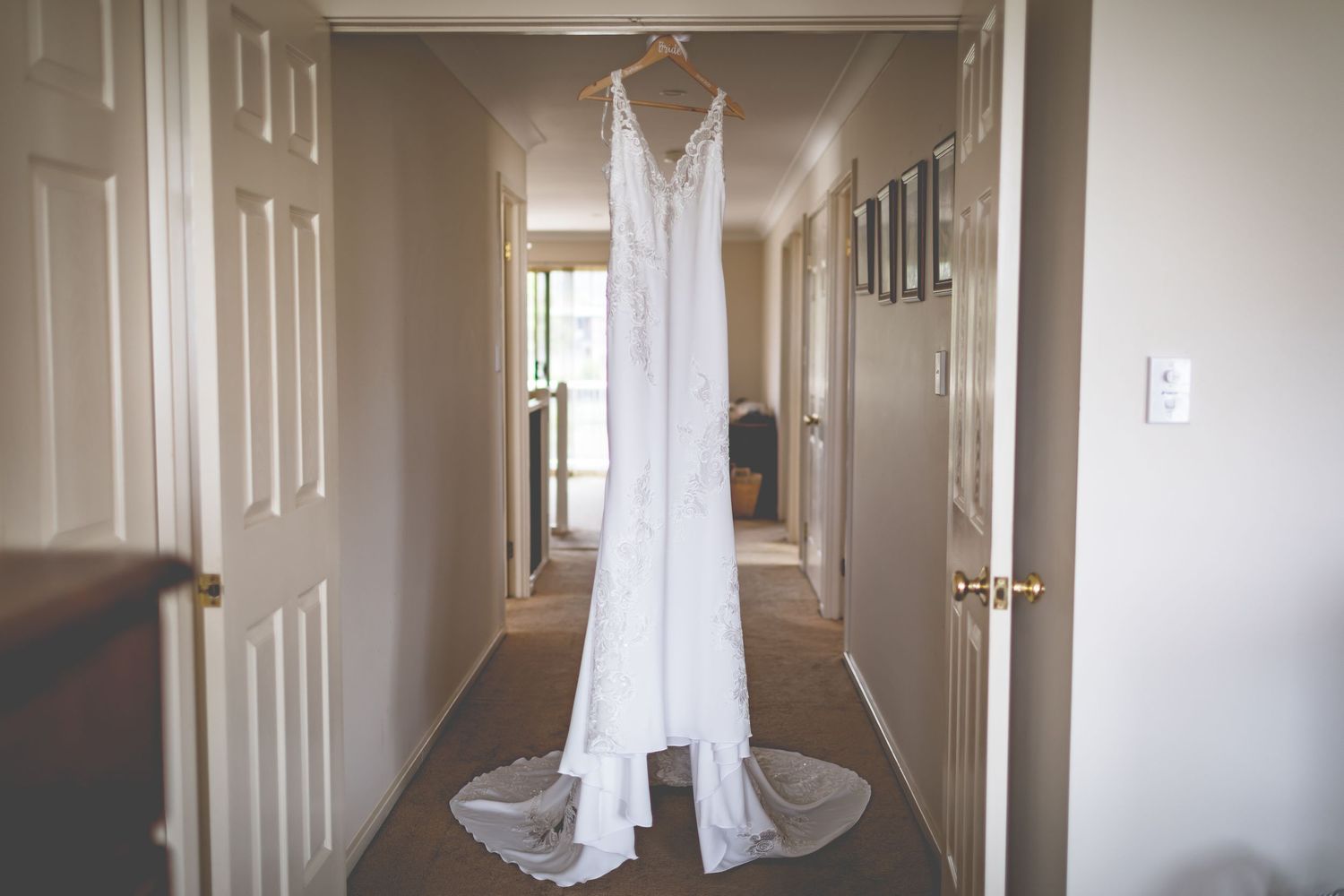 Elegant white lace wedding dress hanging in hallway with natural light streaming through.