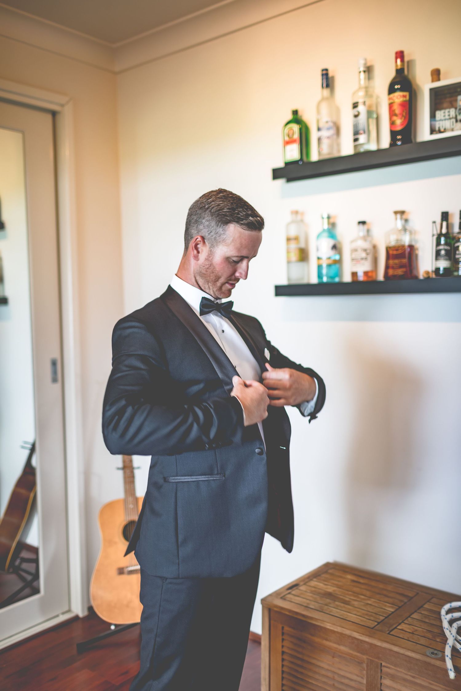 Groom adjusting tuxedo jacket in front of bar shelf with liquor bottles.