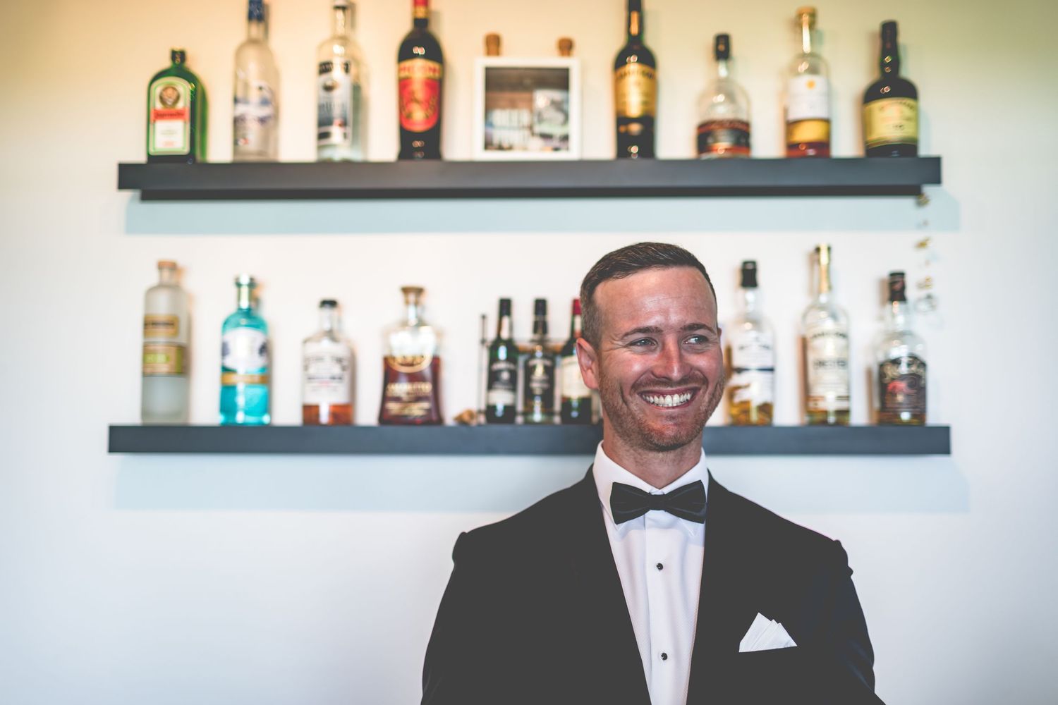 Formal portrait in front of bar shelves displaying various liquor bottles.
