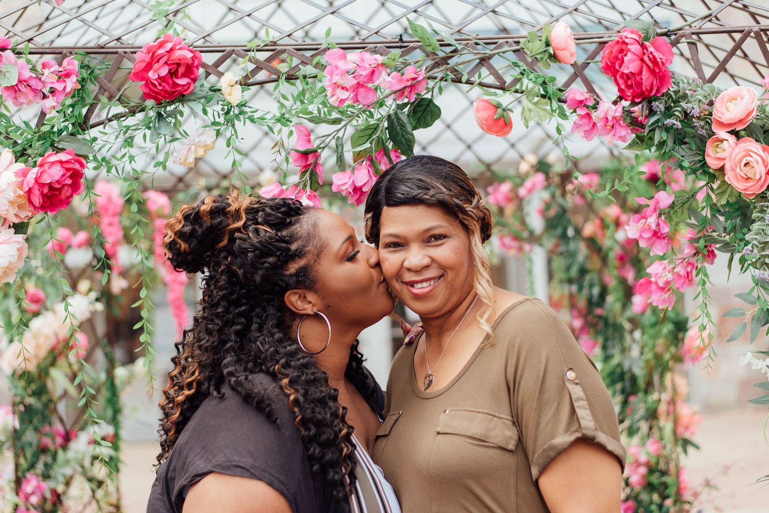 Two people share a sweet moment under a floral arch decorated with pink and coral roses.