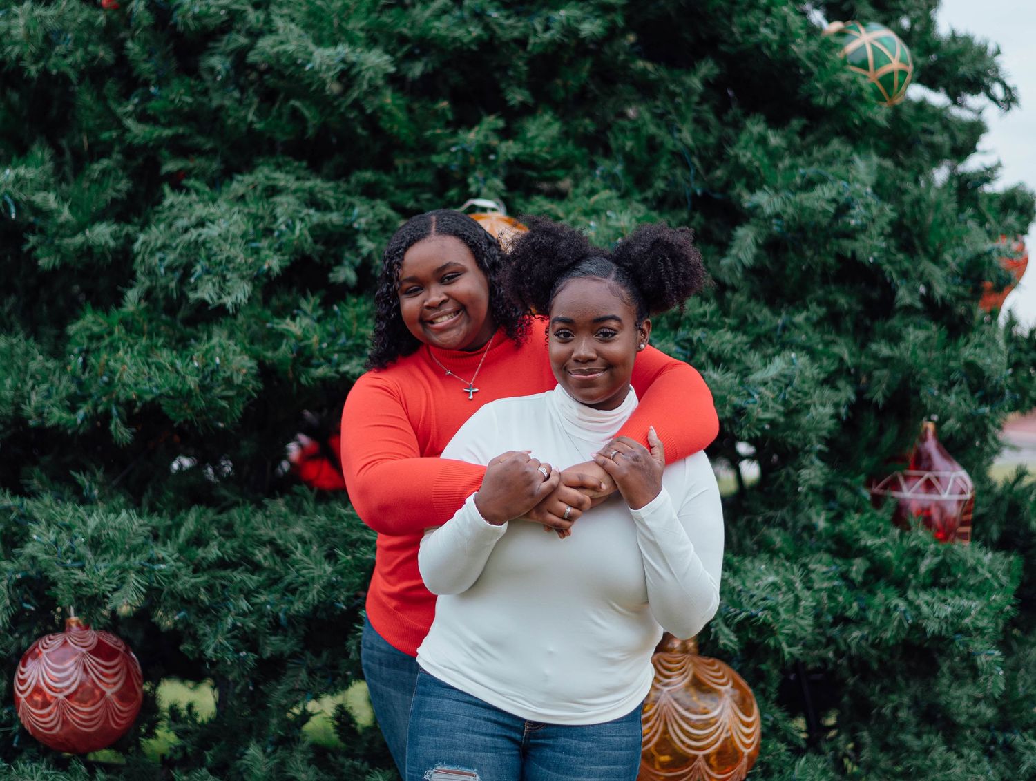Two people embracing and laughing together in front of an evergreen tree with holiday decorations.