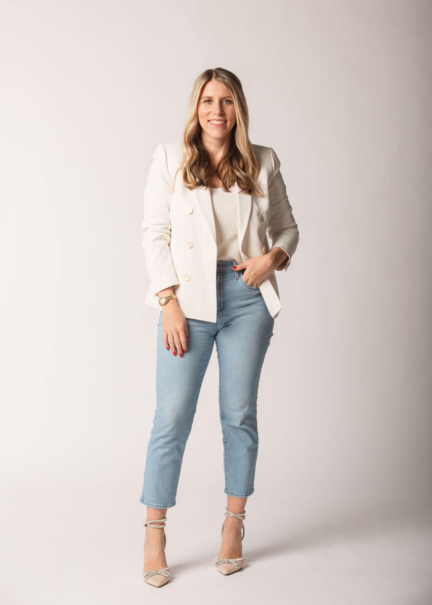 A blonde woman in a white blazer and light blue jeans poses confidently in a professional studio setting.