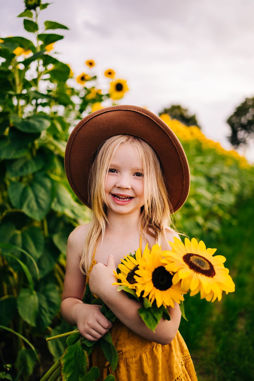 A bright sunflower garden scene with a brown hat and yellow dress creating a cheerful summer portrait.