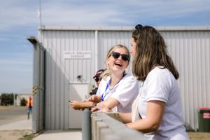 Two people share a laugh outside an aircraft hangar on a sunny day.