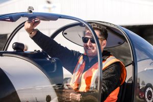 A pilot in safety vest gives thumbs up from aircraft cockpit with jovial expression.