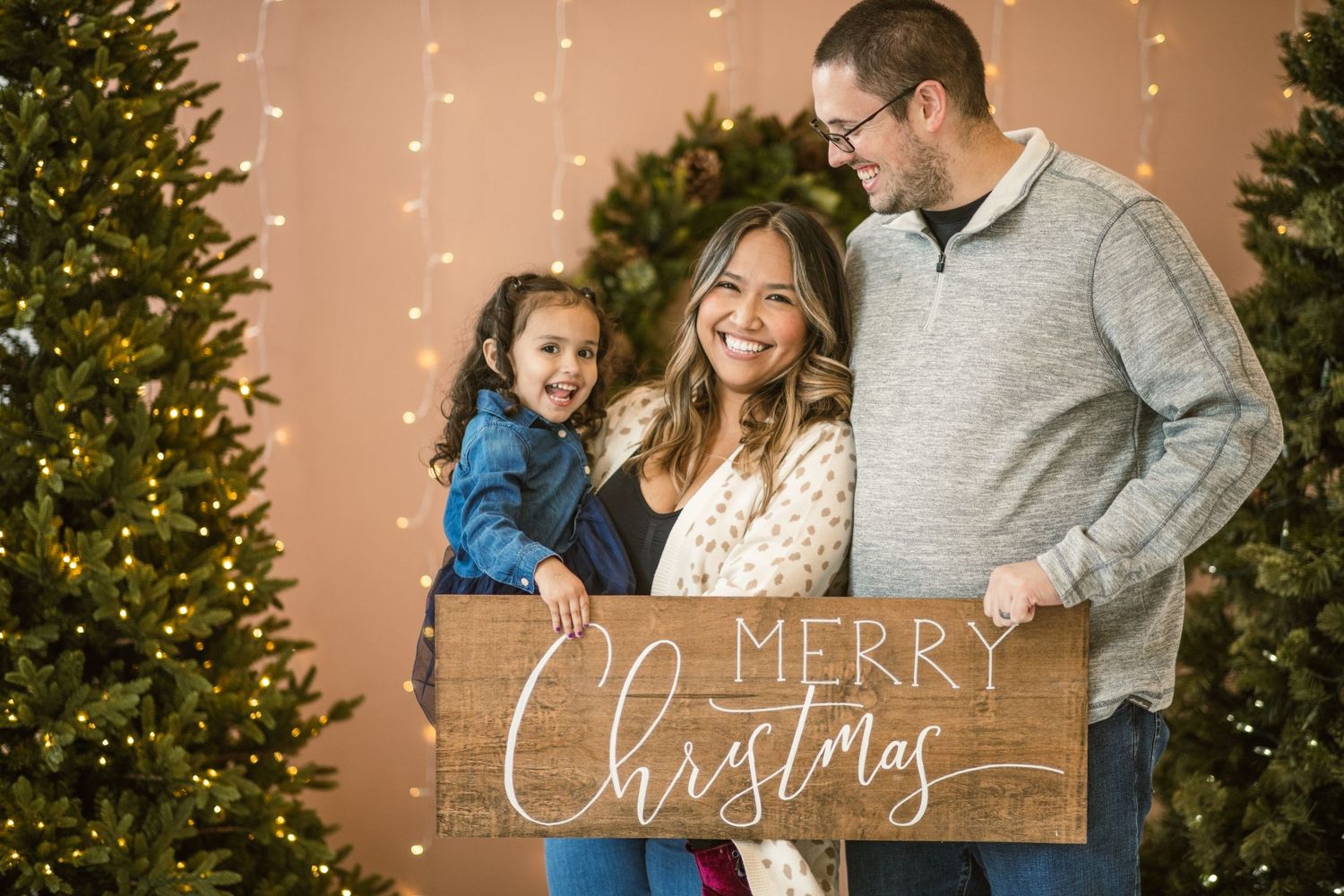 Christmas mini session. Family of 3 holding merry Christmas sign. wife looking at camera. dad looking at daughter.