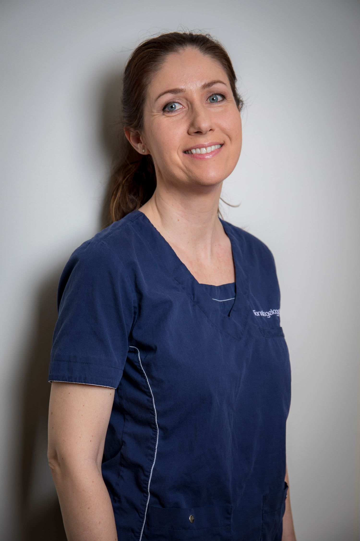Female healthcare professional smiling in navy blue medical scrubs against a light gray background.