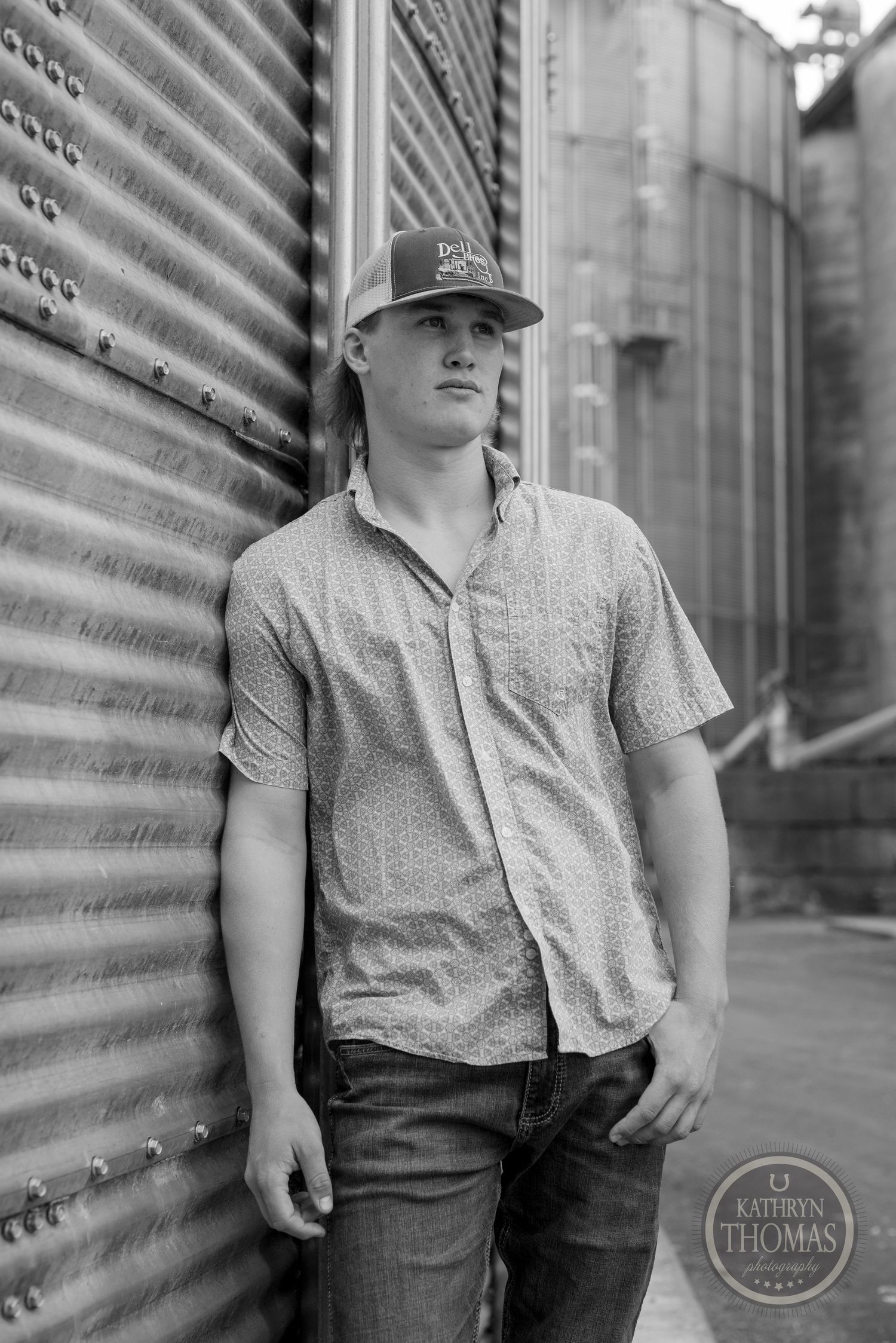 Black and white portrait against corrugated metal siding with grain silos in background.