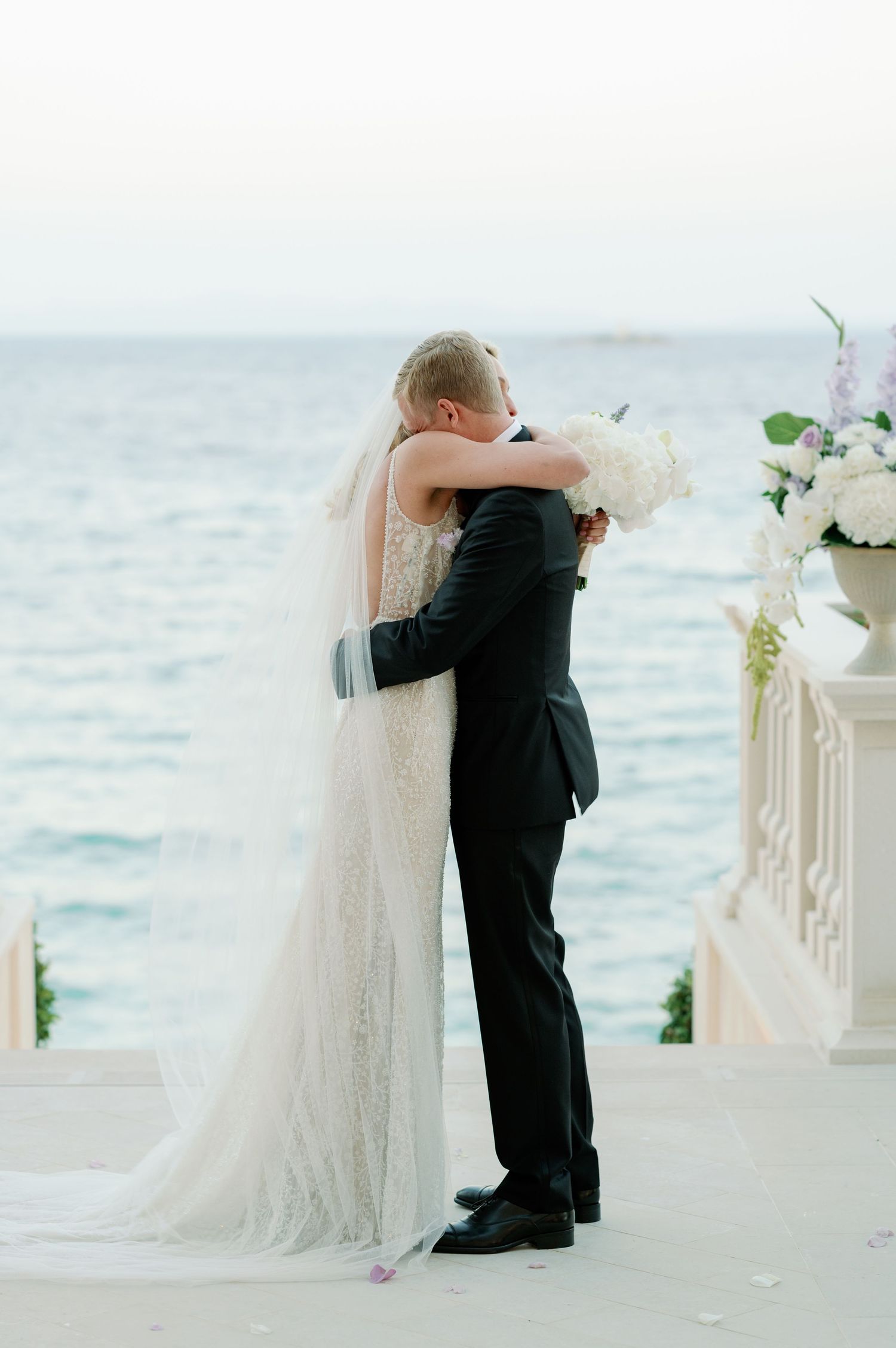 A romantic wedding moment showing a couple embracing on a seaside terrace with white floral decorations.