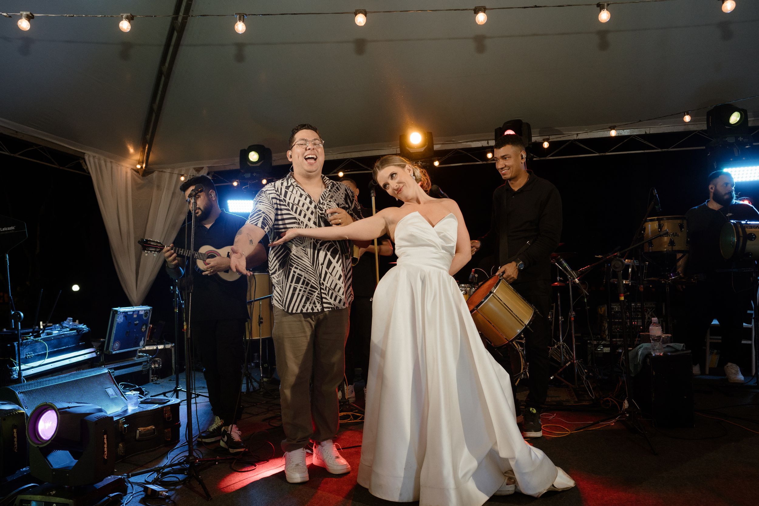 Wedding reception couple dancing on stage with live band performing under string lights at night.