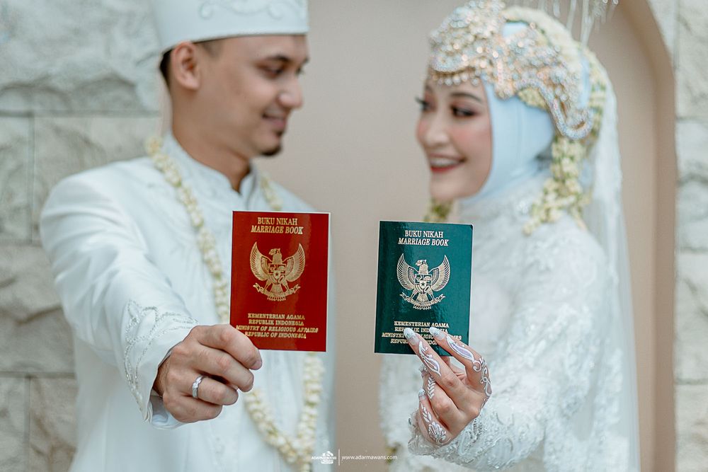 Couple showing Indonesian marriage documents in traditional white and gold wedding attire.