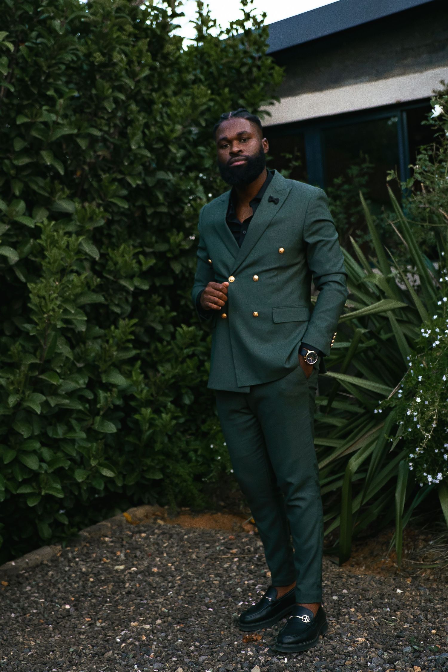 Stylish portrait of someone in a dark green double-breasted suit standing in a garden setting.